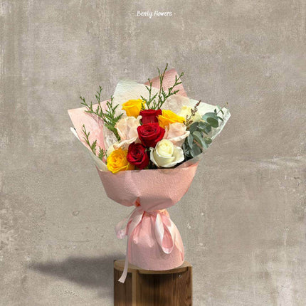 Elegant bouquet featuring vibrant red, yellow, white roses, and lush greenery wrapped in soft pink paper.
