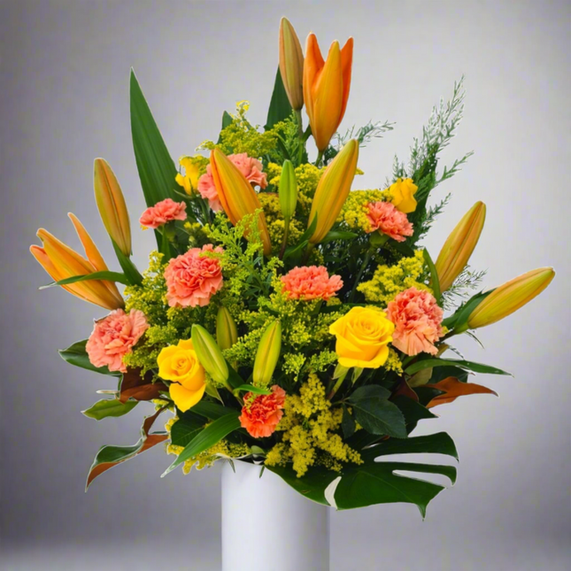 Vibrant flower arrangement with orange lilies, peach carnations, yellow roses, goldenrod, and lush greenery in a white box