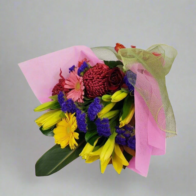 Colorful bouquet featuring roses, lilies, gerberas, and statice, wrapped in pink and green fabric.