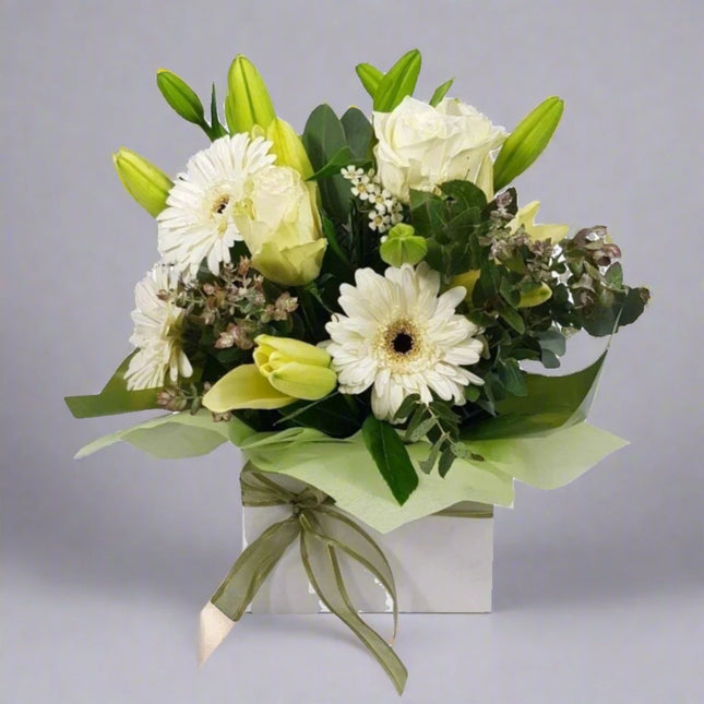 A serene floral arrangement in a white box featuring white lilies, roses, gerbera daisies, and delicate foliage, tied with a green ribbon.