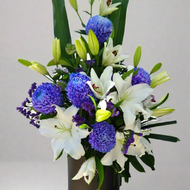 Royal Grace floral arrangement with white lilies, blue chrysanthemums, and purple statice in a black vase.