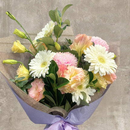 Bouquet of pastel-colored flowers with white daisies, pink chrysanthemums, and yellow roses wrapped in a burlap cover.