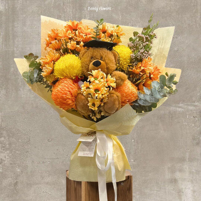 Teddy Flame bouquet featuring orange blooms and a graduation teddy bear, celebrating academic success.