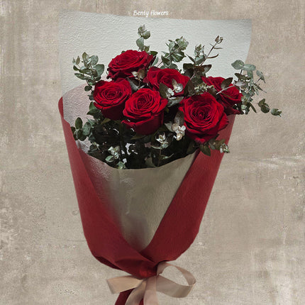 Luxury red rose bouquet wrapped in red and white paper, tied with a beige ribbon, and accented with green foliage. Perfect for romantic gifts, anniversaries, and special occasions – Betty Flowers.