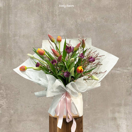 A close-up of a tulip bouquet with a mix of red, yellow, and purple flowers wrapped in white paper.