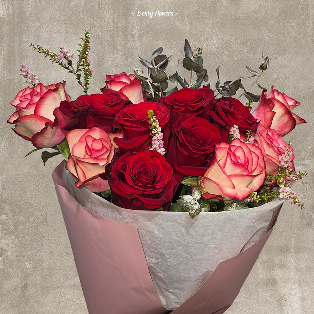 A beautifully arranged bouquet of red and pink roses wrapped in soft pink paper, accented with delicate greenery and small pink flowers. The bouquet has a romantic and elegant feel, ideal for special occasions. The background features a subtle textured surface with the text 'Bently Flowers' at the top.