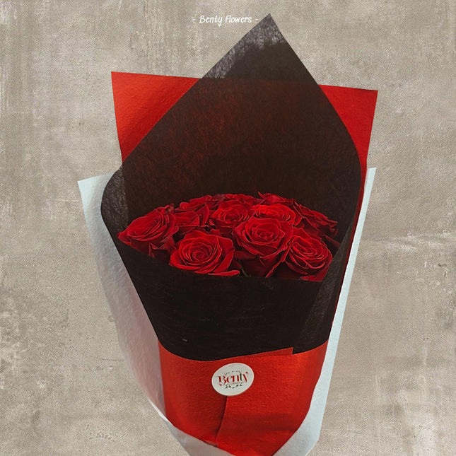 A luxurious bouquet of fresh, deep red roses wrapped in a stylish combination of black, red, and white paper. The bouquet is presented in an elegant and modern design with a 'Bently Flowers' sticker on the front. The roses are arranged in a tight, circular pattern, creating a striking and romantic aesthetic.