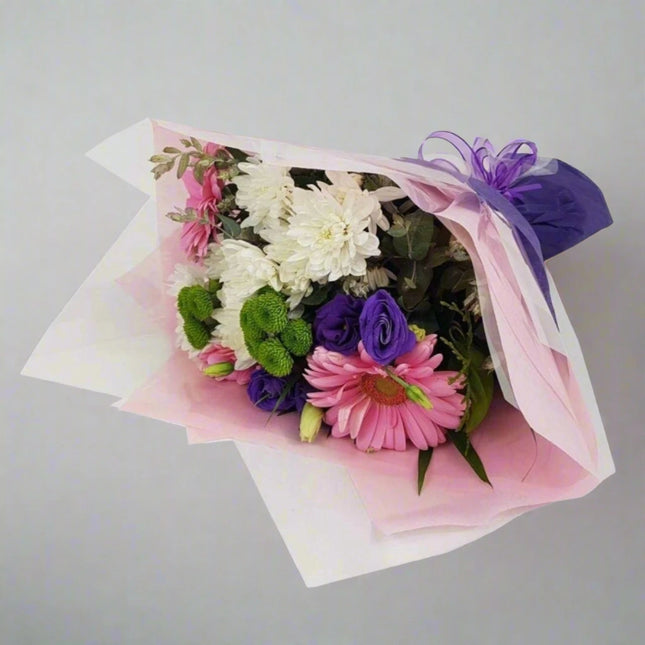 Blissful Blooms Bouquet featuring white chrysanthemums, pink gerbera daisies, purple lisianthus, and green button poms, elegantly wrapped in pink and white paper with a purple ribbon.