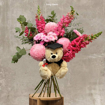 A delightful pink graduation bouquet featuring pink snapdragons, chrysanthemums, and a charming teddy bear dressed in a graduation cap, wrapped in soft white paper with a pink ribbon. Perfect for celebrating academic milestones with elegance.