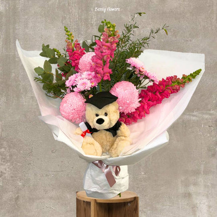 A delightful pink graduation bouquet featuring pink snapdragons, chrysanthemums, and a charming teddy bear dressed in a graduation cap, wrapped in soft white paper with a pink ribbon. Perfect for celebrating academic milestones with elegance.