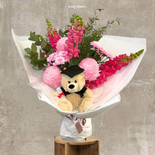 Pink graduation bouquet with snapdragons, chrysanthemums, and a teddy bear in a cap, celebrating graduates.