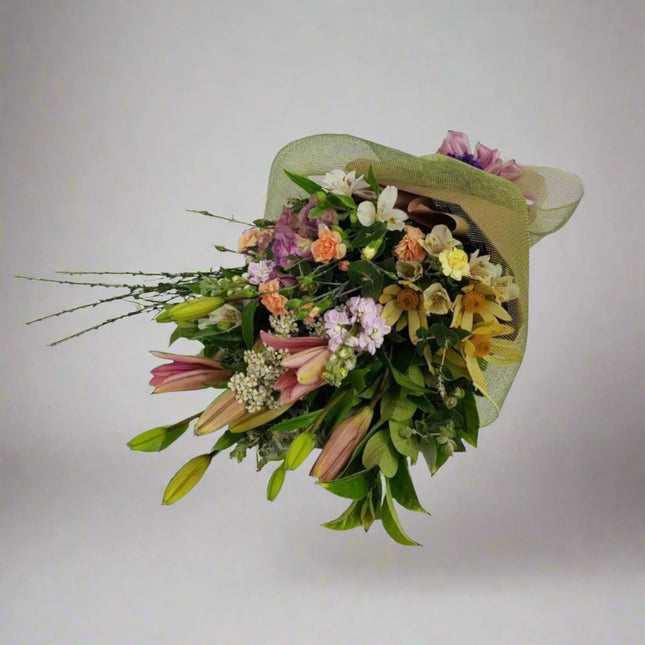 A beautifully arranged hand-tied bouquet featuring a variety of delicate flowers including lilies, roses, and seasonal blooms, wrapped in a soft green mesh.