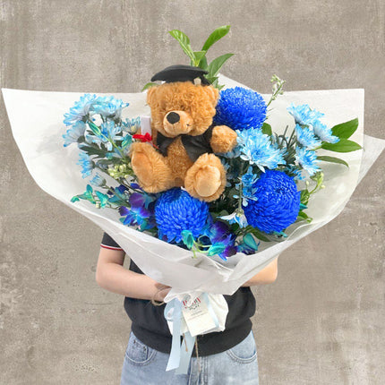 A vibrant blue graduation bouquet featuring blue chrysanthemums, delicate floral accents, and a lovable teddy bear dressed in a graduation cap, beautifully wrapped in white paper. Ideal for celebrating academic achievements with charm and style.