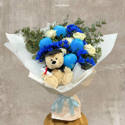 Graduation bouquet featuring elegant blue chrysanthemums, white roses, lush greenery, and a charming teddy bear in a graduation cap, celebrating academic success with style and grace.