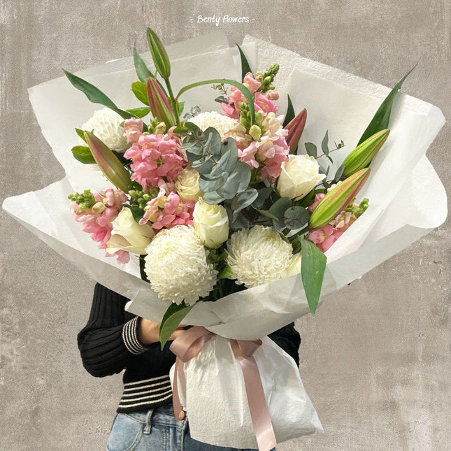 A stunning graduation bouquet showcasing elegant oriental lilies, soft pink snapdragons, creamy white roses, and fluffy chrysanthemums. This lush arrangement is complemented by fresh eucalyptus leaves and wrapped in crisp white paper with a delicate pink ribbon, embodying sophistication and celebration.