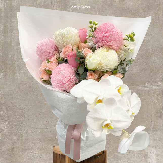 A sophisticated graduation bouquet featuring elegant white orchids, soft pink and white chrysanthemums, pastel roses, and lush greenery. This delicate arrangement is wrapped in pristine white paper with a satin pink ribbon, perfect for celebrating academic achievements with grace and beauty.