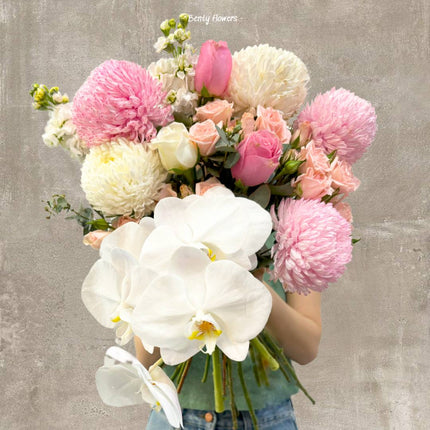 A sophisticated graduation bouquet featuring elegant white orchids, soft pink and white chrysanthemums, pastel roses, and lush greenery. This delicate arrangement is wrapped in pristine white paper with a satin pink ribbon, perfect for celebrating academic achievements with grace and beauty.