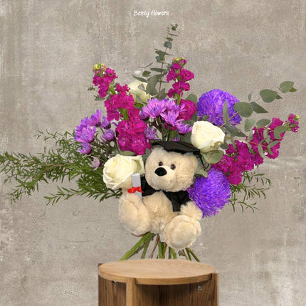 A stunning purple-themed graduation bouquet showcasing vibrant purple flowers, elegant white roses, and lush greenery, accented with a charming teddy bear in a graduation cap. Perfect for celebrating academic milestones with sophistication and joy.