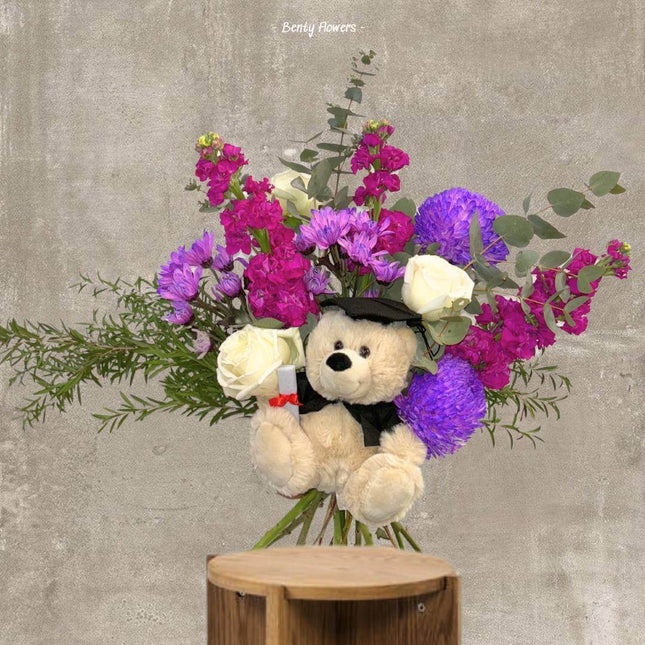 Purple graduation bouquet with flowers, greenery, and a cute teddy bear in a graduation cap.