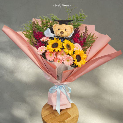 A vibrant graduation bouquet featuring sunny sunflowers, cheerful pink gerberas, and a mix of lush greenery, beautifully wrapped in pink paper. Highlighted with a cute teddy bear wearing a graduation cap, this arrangement radiates celebration and joy.